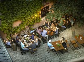 Menschen sitzen an Holztischen und essen; sie unterhalten sich unter Lichterketten.