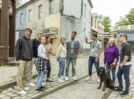 A diverse group of smiling teenagers and a dog pose together on a cobbled street.
