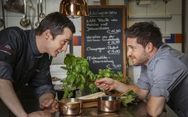 Zwei Mitarbeiter der Küche stehen sich in Kochjacken in der Küche gegenüber und blicken zueinander. Mittig im Bildhintergrund hängt die aktuelle Speisekarte des Restaurants Pfistermühle an der Wand.