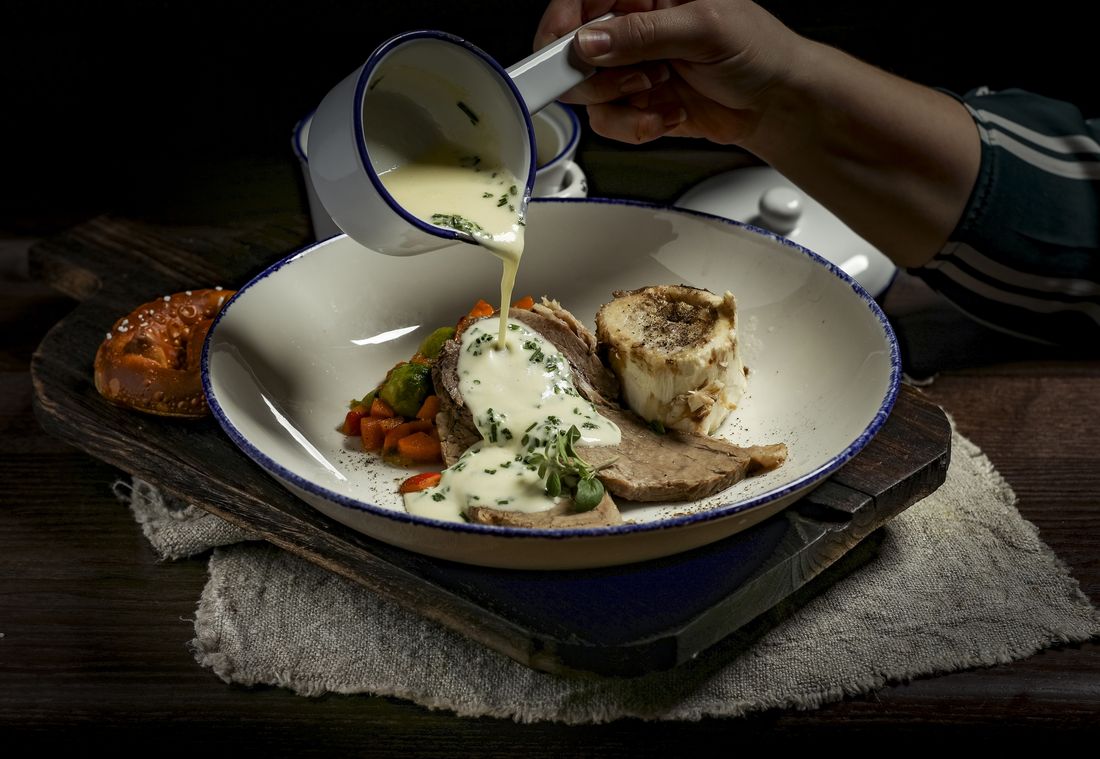 A hand pours creamy sauce from a small pitcher onto slices of roasted meat alongside vegetables in a white bowl.