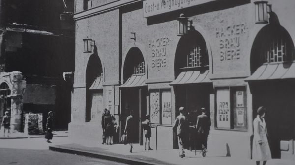 A historical picture of a building with the inscription "Hackerbräu Biere", in front of which several passers-by walk