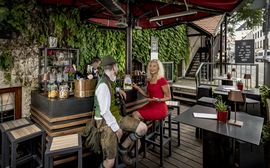A man in Bavarian costume sits with a blonde lady in a red dress at the bar in Platzl Karree.
