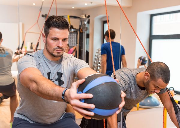Zwei Männer konzentrieren sich auf ein Krafttraining mit einem Gewichtsball in einem hellen Fitnessraum mit Spiegeln und Widerstandsbändern