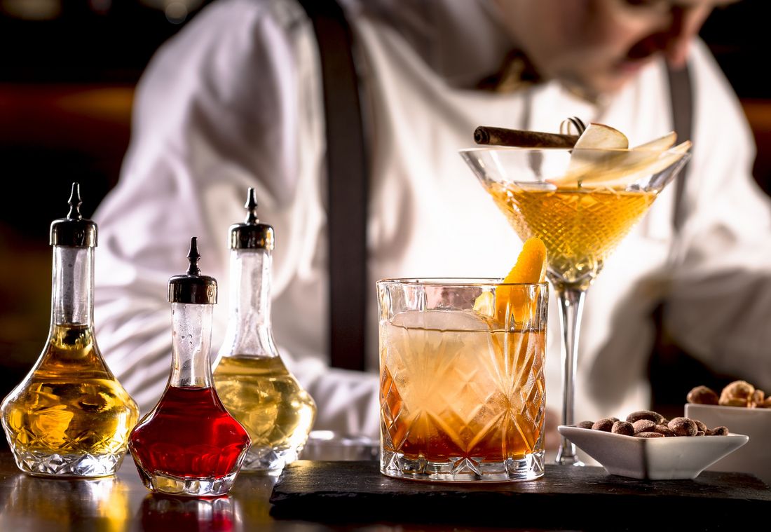 A glass with an orange slice stands next to a cocktail glass, with a bartender working in the background.