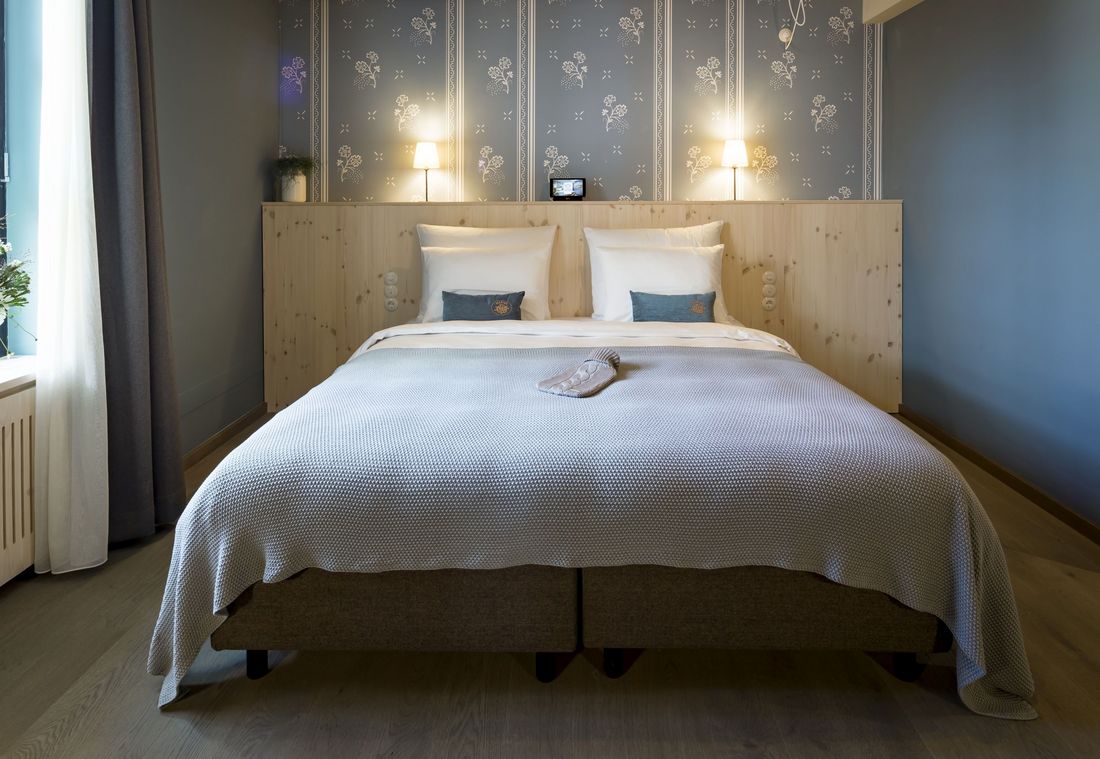 Bed with white bedspread and pillows; above it wall lamps and wallpaper with a floral pattern in the background;