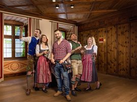 A team dressed in dirndls and lederhosen stands with a craftsman in a rustic wooden room.