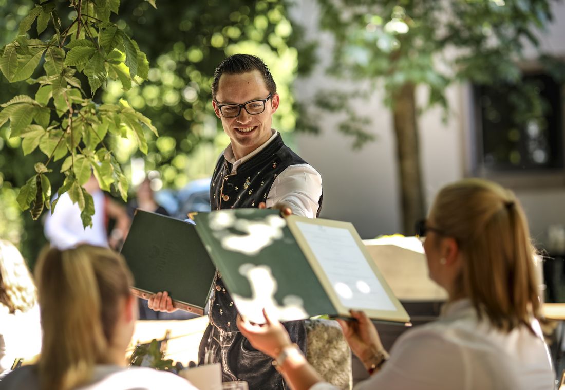 A person in traditional attitude smiles as holding a green folder outdoors, amidst leafy trees and dappled.