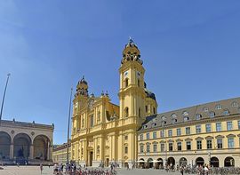 Die gelbe Theatinerkirche am Odeonsplatz in München vor einem blauen Himmel mit Passanten.