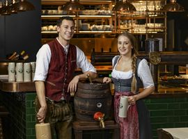Zwei Mitarbeiter des Ayinger in der Au stehen zusammen in Tracht an der Bar