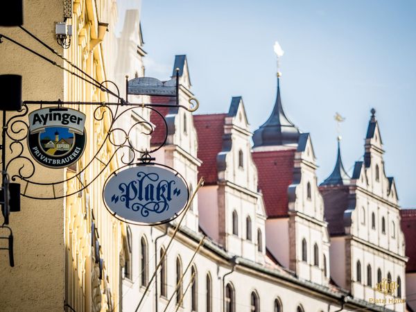 A view along the facades of houses in Munich, on which the logo of the Ayinger brewery and a logo of the Platzl Hotel Munich hang