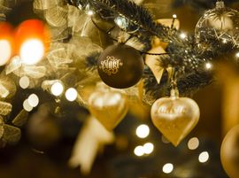 A close-up of a decorated Christmas tree with gold ornaments, including a heart-shaped ornament and blurred lights in the background.