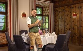 Ein Mitarbeiter der Platzl Hotels steht in bayerischer Tracht vor einem Tisch mit grauen Stühlen.