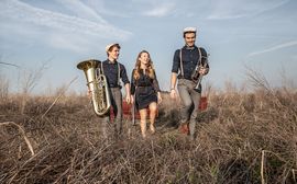 Three young musicians of the brass band "DIE FEXER" are standing in a field with their instruments.