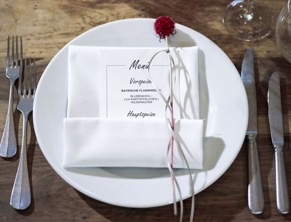 Detailansicht eines Tischgedecks in der Hochzeitslocation Marias Platzl mit Gabeln, Messern und einem Teller, auf dem mittig eine Menükarte mit roter Blume als Dekoration platziert wurde.