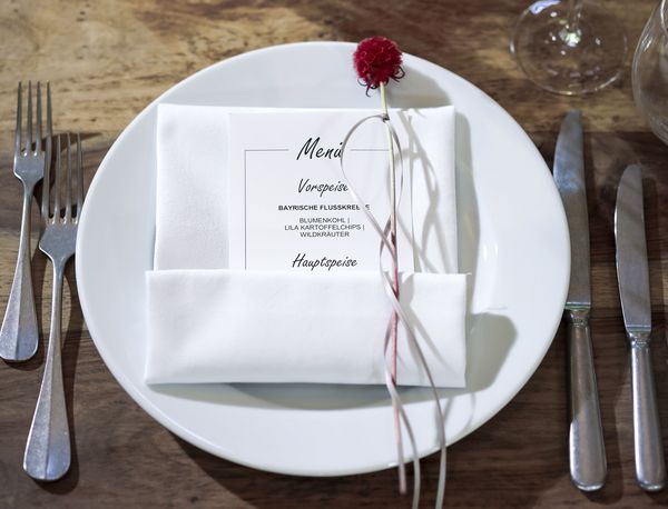 Platzteller auf einem gedeckten Tisch in der Event Location in München Detailansicht eines Tischgedecks in der Hochzeitslocation Marias Platzl mit Gabeln, Messern und einem Teller, auf dem mittig eine Menükarte mit roter Blume als Dekoration platziert wurde.