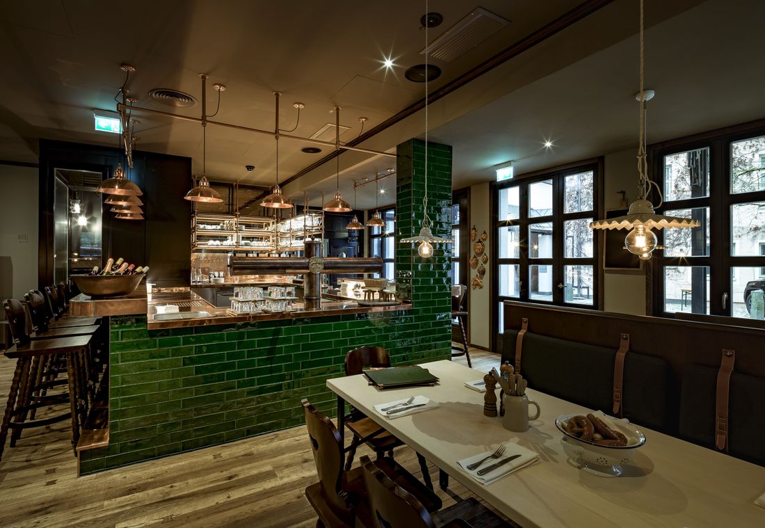 A restaurant interior with wooden tables and chairs; a counter with green tiles dominates the center.